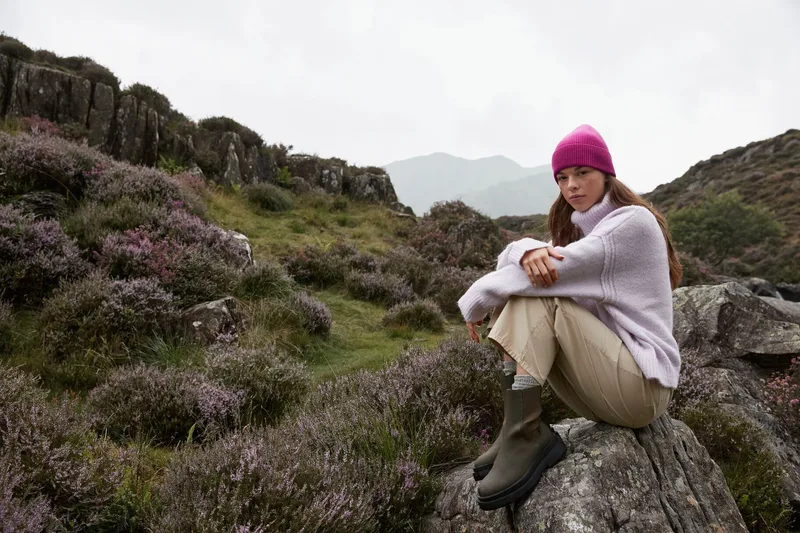 Frau mit pinker Dip-Dye-Beanie sitzt auf Felsen in Hügellandschaft
