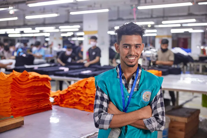 Junger Arbeiter mit verschränkten Armen in einer Fabrikhalle, neben ihm Stapel oranger Textilien. Bild aus dem Artikel zur Fair-Wear-Mitgliedschaft.