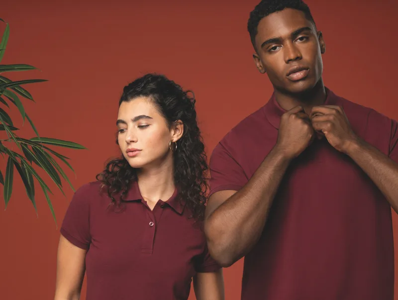 Frau und Mann in weinroten Poloshirts vor einer roten Wand mit Zimmerpflanze. Titelbild der Organic-Kollektion.