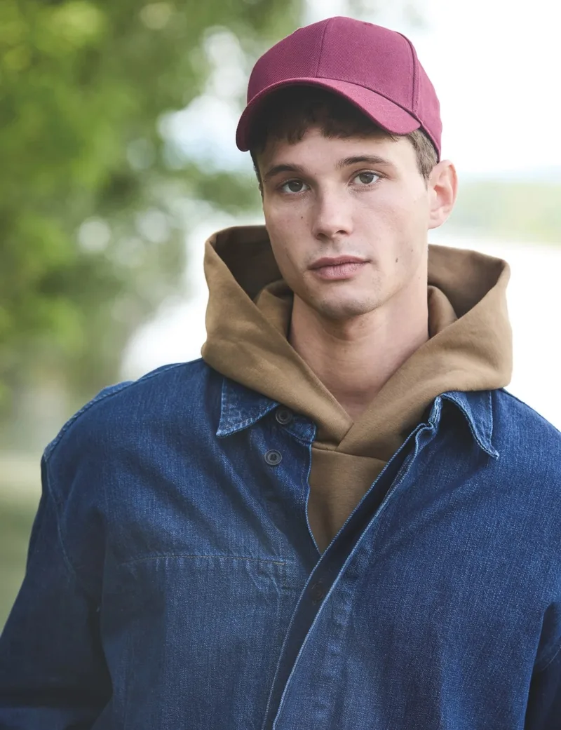 Junger Mann mit weinroter Kappe, Jeansjacke und Kapuzenpulli draußen vor Bäumen. Kollektion Gold Label. Hochformat.