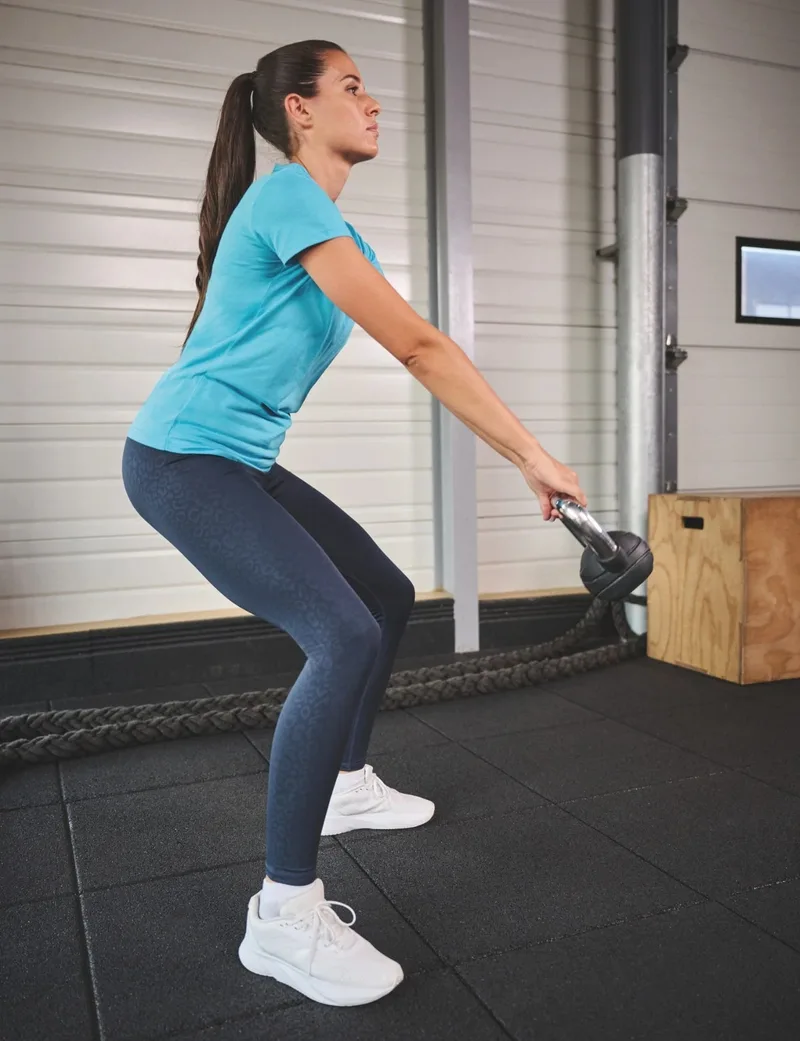 Frau in türkisem Shirt beim Kettlebell-Training in einem Fitnessraum. Hochformat.