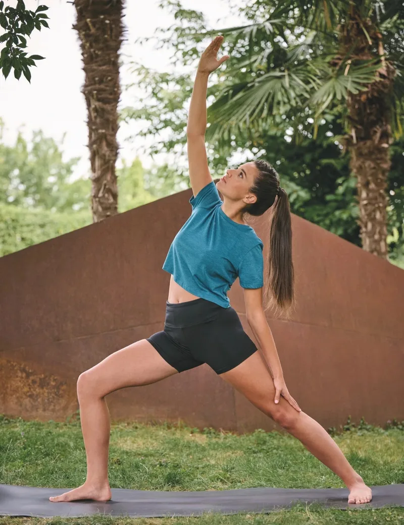 Frau in türkisem Shirt macht draußen auf einer Yogamatte eine Yoga-Pose, im Hintergrund Palmen und eine Mauer. Hochformat.