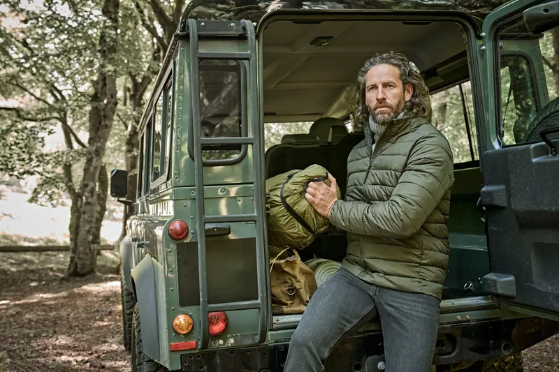 Mann in Steppjacke sitzt an einem alten grünen Geländewagen im Wald (ca. 34 MB)
