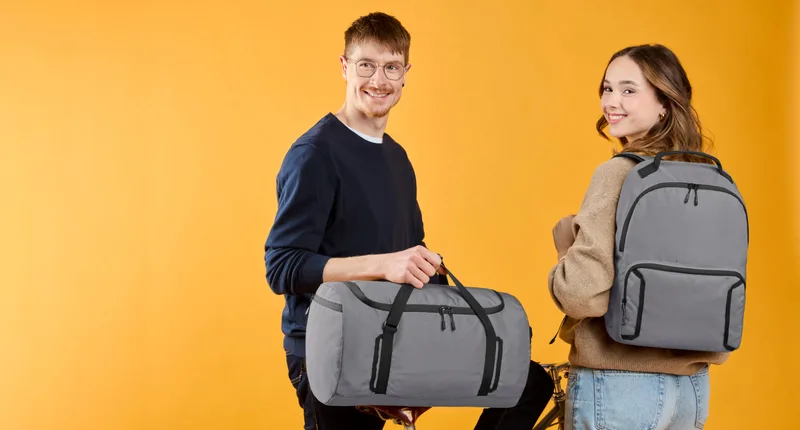 Junger Mann und junge Frau mit grauer Tasche und grauem Rucksack der Serie LOOP vor orange-gelbem Studiohintergrund (Datei ca. 27 MB)