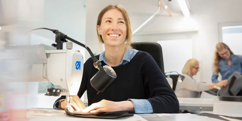 Lächelnde Näherin an der Nähmaschine, im Hintergrund weitere Kolleginnen an ihren Plätzen (Ausbildung Textil- und Modenäher)