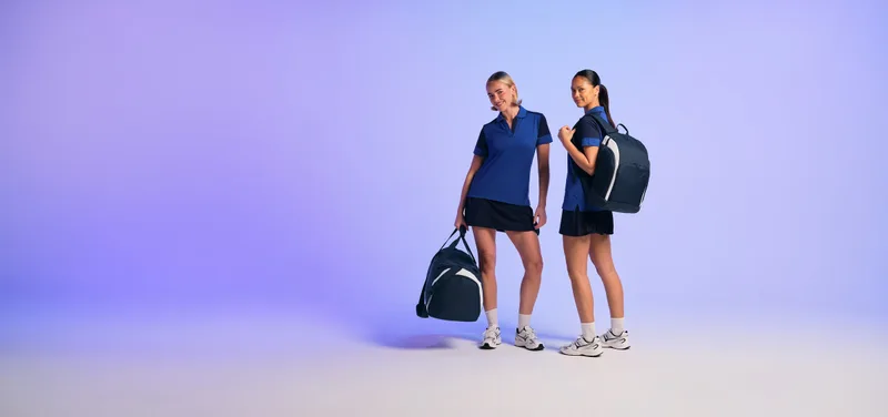 Zwei Sportlerinnen in navyblauer Teamkleidung mit Teamwear-Rucksack und Sporttasche vor lila Studiohintergrund