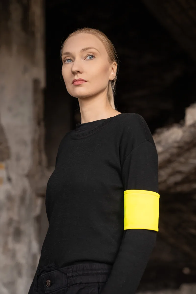 Frau im schwarzen Pullover mit gelber Reflex-Armbinde vor rauer Betonwand (Hochformat, ca. 9,5 MB)