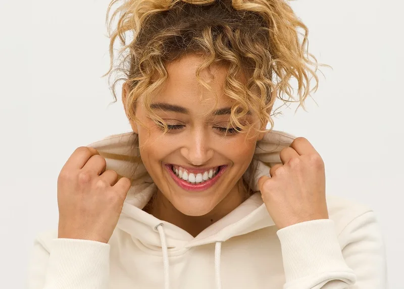 Lachende Frau mit Locken zieht sich die Kapuze eines cremefarbenen Hoodies ins Gesicht, heller Studiohintergrund.