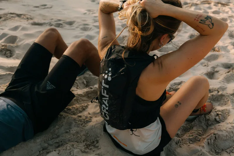 Zwei Trailläufer in Craft-Laufweste und Shorts ruhen sich im Sand aus (Rückenansicht, Craft-Schriftzug auf der Weste)