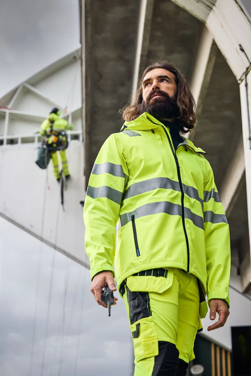 Bärtiger Arbeiter in gelber Projob-Warnschutzjacke vor einem Rohbau, ein Kollege im Hintergrund auf dem Gerüst (Hochformat)