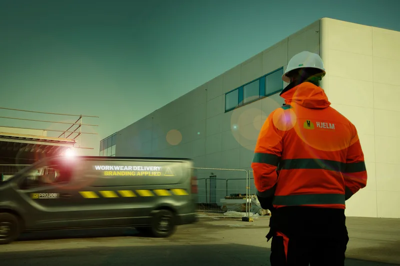 Mann in orangefarbener Projob-Warnschutzjacke vor einem Industriegebäude, im Hintergrund ein Lieferwagen mit Licht (Abendstimmung)