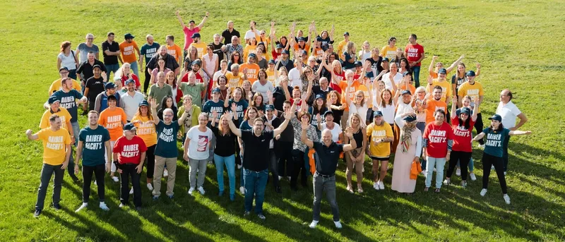 Gruppenfoto der Belegschaft auf einer Wiese, alle in bunten Firmen-Shirts (laut Alt-Text bei einer Unternehmensfeier)